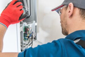 Technician in grey cap and blue t-shirt is repairing a furnace unit.